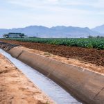 Canal de irrigação atravessando área agrícola, com montanhas ao fundo.