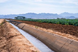 Canal de irrigação atravessando área agrícola, com montanhas ao fundo.