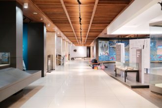 Corredor amplo e iluminado de uma exposição de design contemporâneo, com obras dispostas ao longo do espaço.