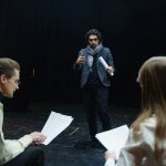 Atores e diretor analisam roteiros durante ensaio em um teatro.