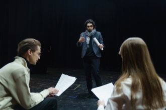 Atores e diretor analisam roteiros durante ensaio em um teatro.