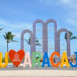 Placa com a inscrição “EU AMO ARACAJU” em uma área ao ar livre, cercada por palmeiras sob céu azul.