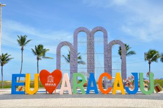 Placa com a inscrição “EU AMO ARACAJU” em uma área ao ar livre, cercada por palmeiras sob céu azul.