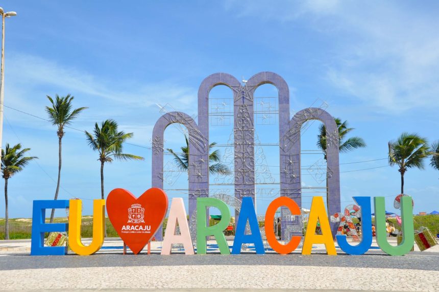 Placa com a inscrição “EU AMO ARACAJU” em uma área ao ar livre, cercada por palmeiras sob céu azul.
